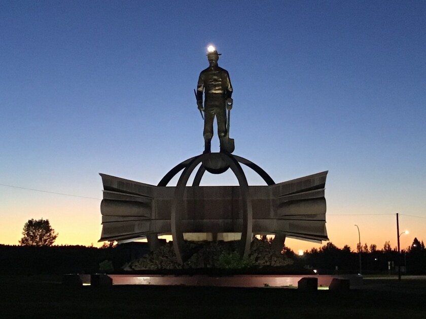 A monumental sculpture of an iron miner standing atop a flared pile of steel girders is seen on a Midwestern landscape at dusk.