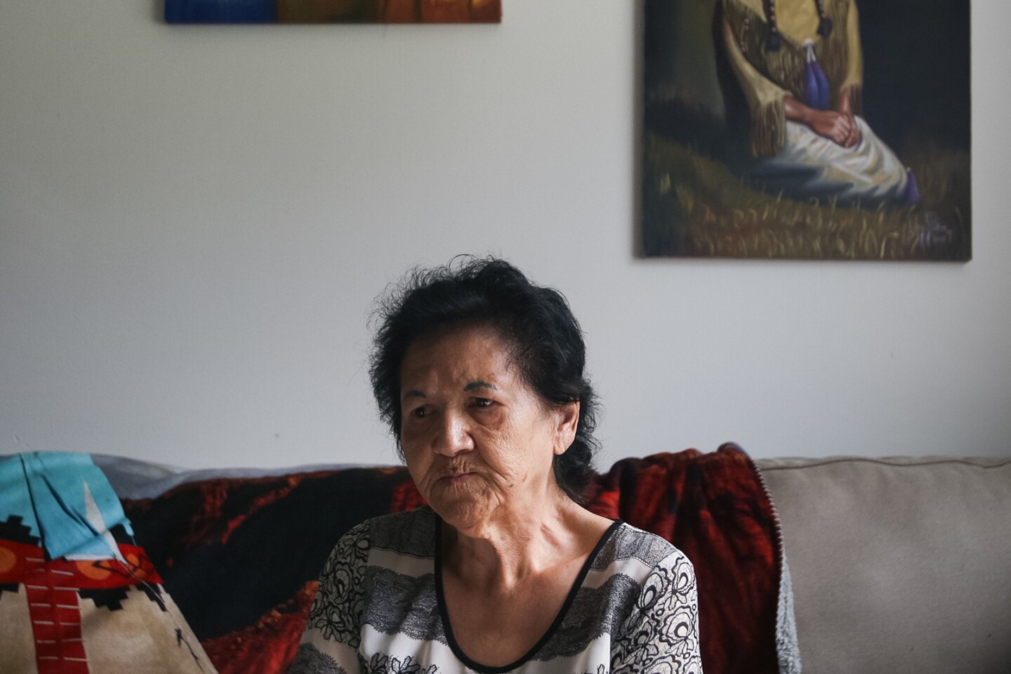 A wizened, dark-haired woman purses her lips while sitting on a couch. Behind her, canvases painted with Indigenous people hang on the wall.