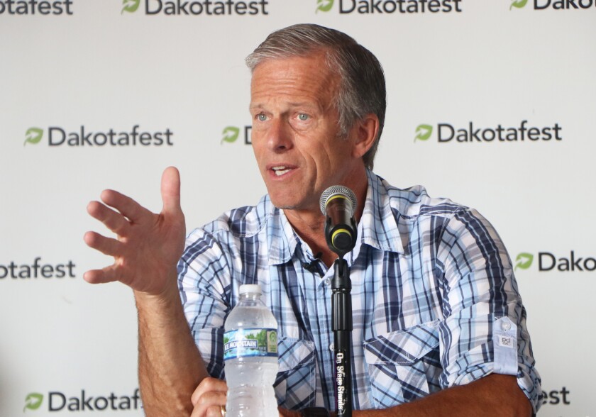 A man in a blue plaid, short-sleeved shirt sits flanked by a Dakotafest farm show backdrop, and speaking into a microphone.