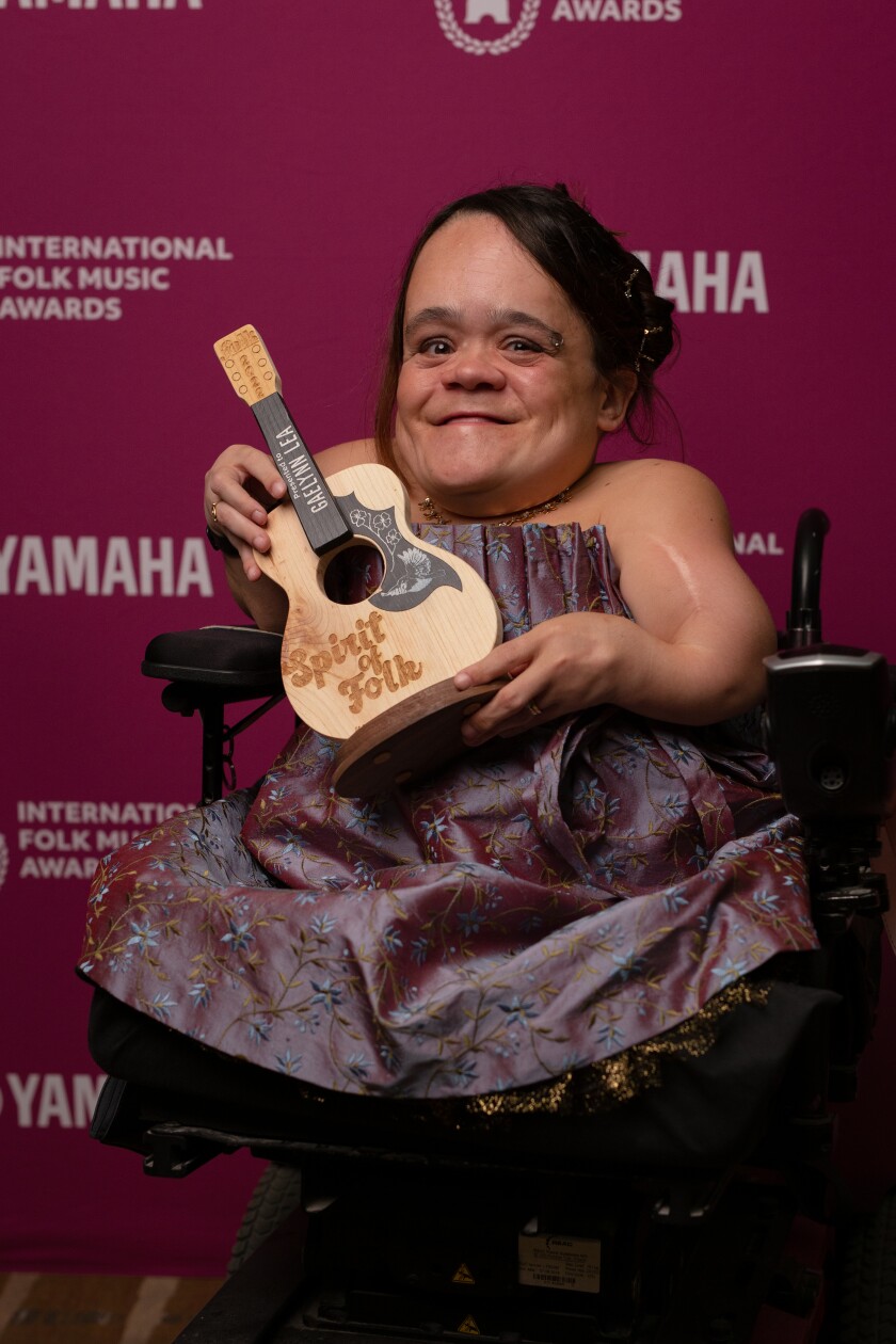 Gaelynn Lea, a white woman wearing a formal dress and sitting in a wheelchair, smiles and poses with a guitar-shaped "Spirit of Folk" award.