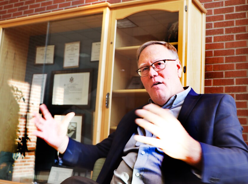 A scientist in a blue sport coat, flanked by trophy cases in a university hallway, gestures with his hands about the seriousness of failing to confront a weed threat.