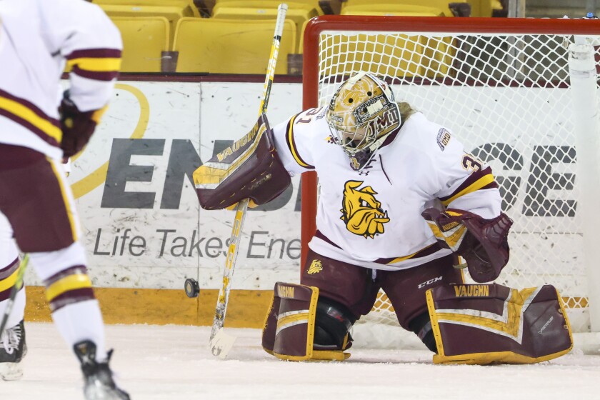 Women hockey players in purple and yellow uniforms compete against players in white and maroon uniforms on an indoor ice sheet.
