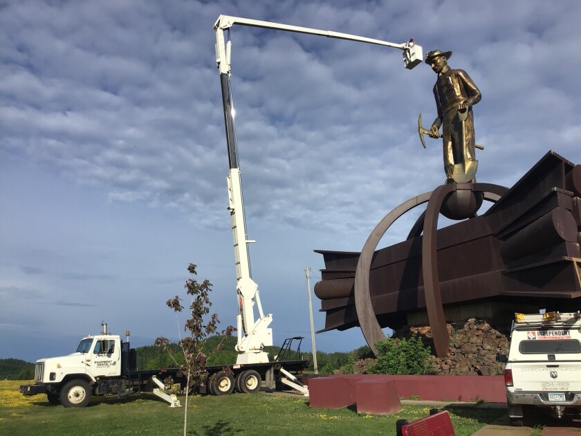 White truck with bucket at end of long crane is seen extending bucket to the front of golden statue of miner standing atop steel beams.