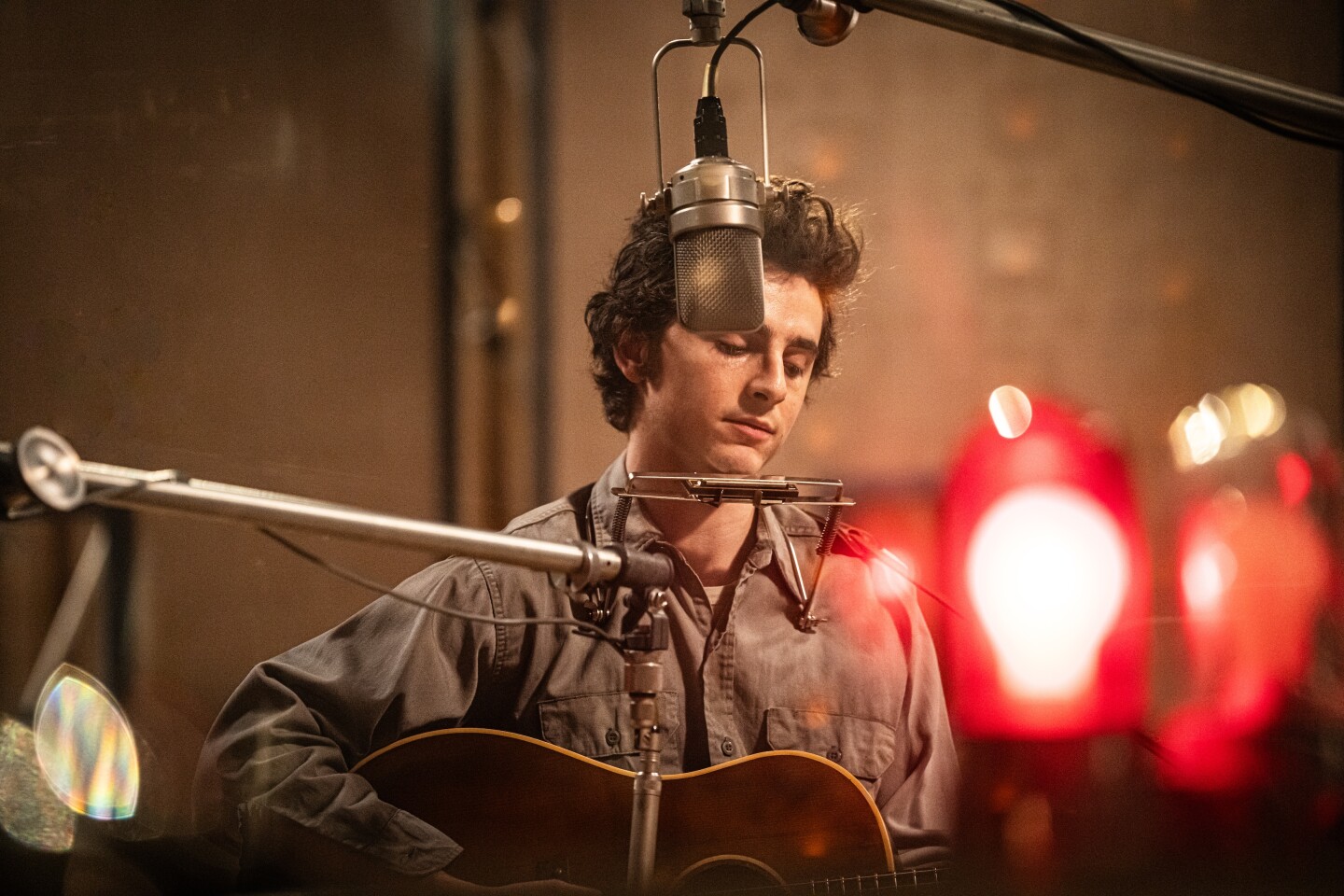 White man in his 20s plays acoustic guitar in a recording studio, with a red recording light seen out-of-focus in the foreground.
