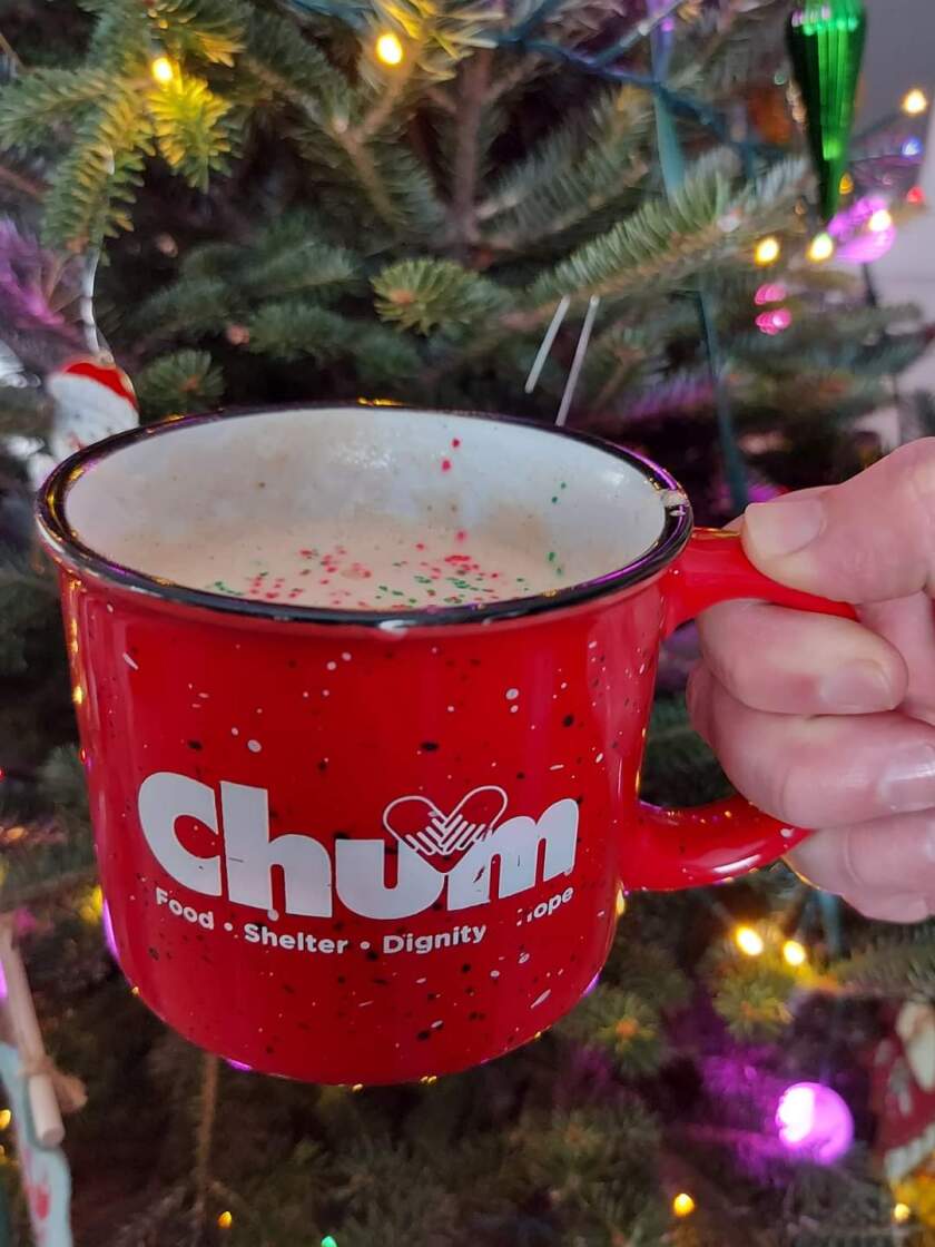 A red mug with the CHUM logo is held by a hand in front of an out-of-focus Christmas tree. A white liquid topped with rainbow sprinkles is visible inside.