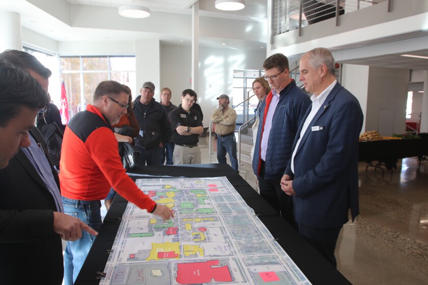 About a dozen people look at a large blueprint while one man gestures to the center of it.