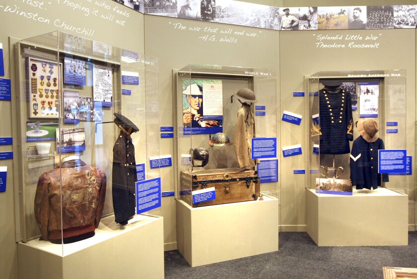 Three display cases in a museum setting include clothing and other artifacts worn during the Spanish American War, World War I and World War II.