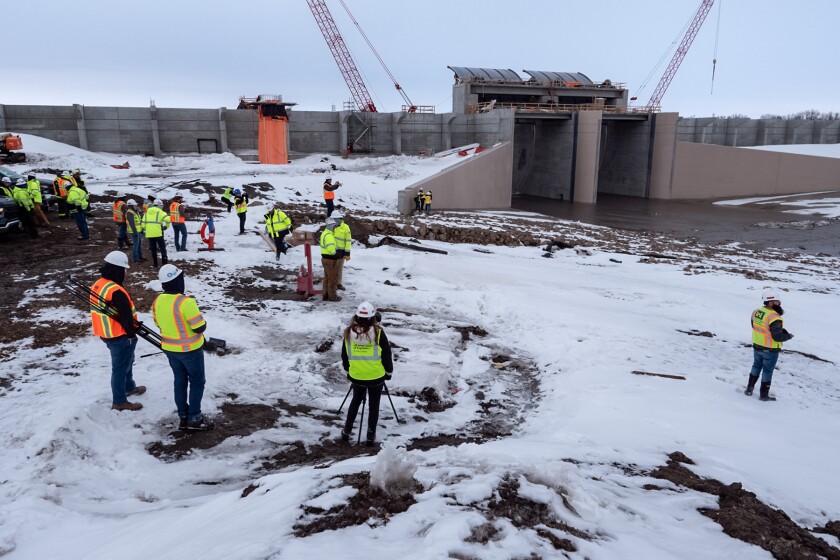 On a snowy, muddy landscape, about 20 people in bright yellow and orange safety vests are dwarfed by a looming concrete structure through which water is flowing.