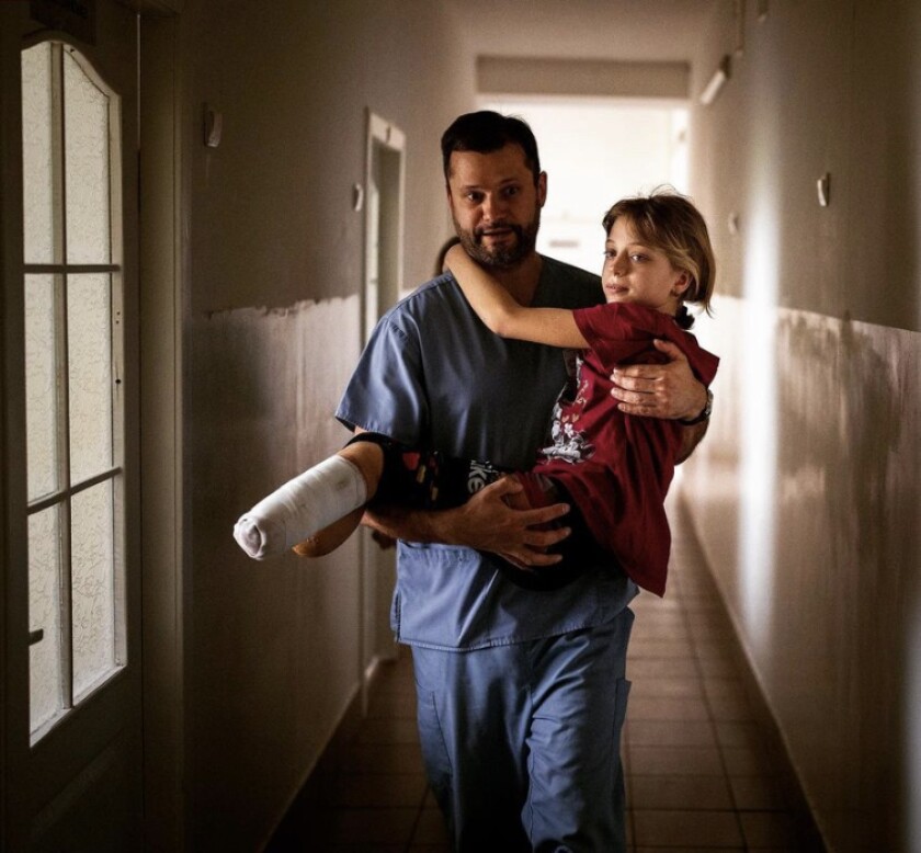 An adolescent girl who is missing the ends of both legs is carried down a hall by a man in scrubs.