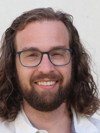 A young man with dark, long hair, beard and glasses, smiles, with a white backdrop.