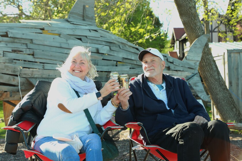 White couple of senior age sit on camp chairs in front of a large outdoor wooden shark sculpture, toasting with two cans of beer.