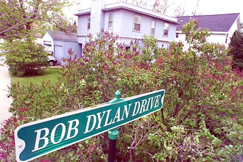 Street sign reading "Bob Dylan Drive" stands in front of a hedge. Behind the hedge, a light-colored, square two-story home is visible across a lawn.