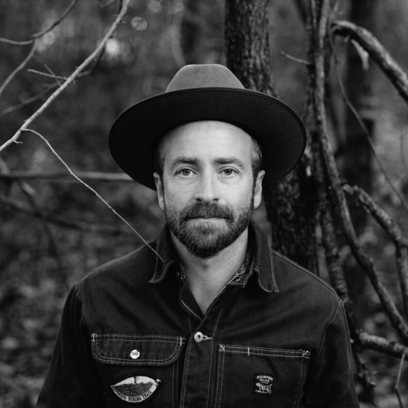 Black and white image of Dave Simonett standing in woods, wearing medium-brim hat and denim-like shirt.