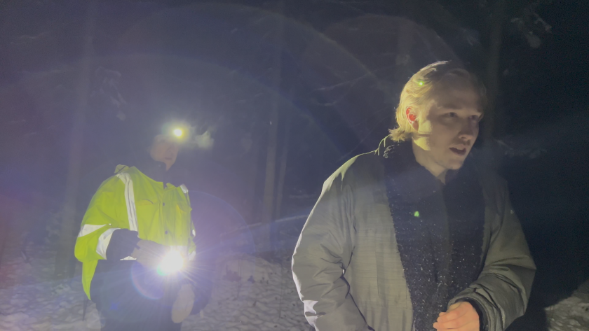 At night in a snowy forest, a man walks purposefully to the right while another man, wearing a reflective vest, follows him with a light.