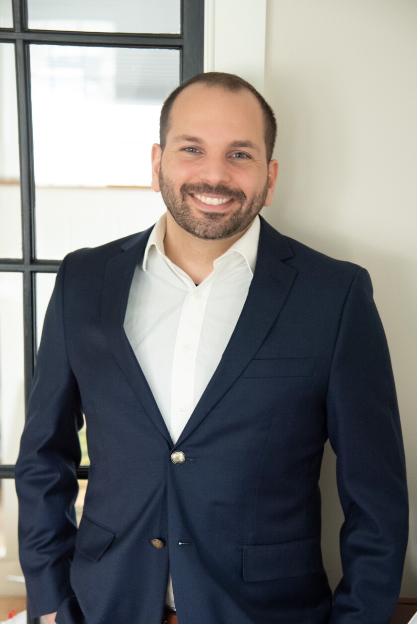 Light-skinned man with dark hair and beard stands smiling for portrait in front of pane-glass window. He wears navy blazer and white shirt.