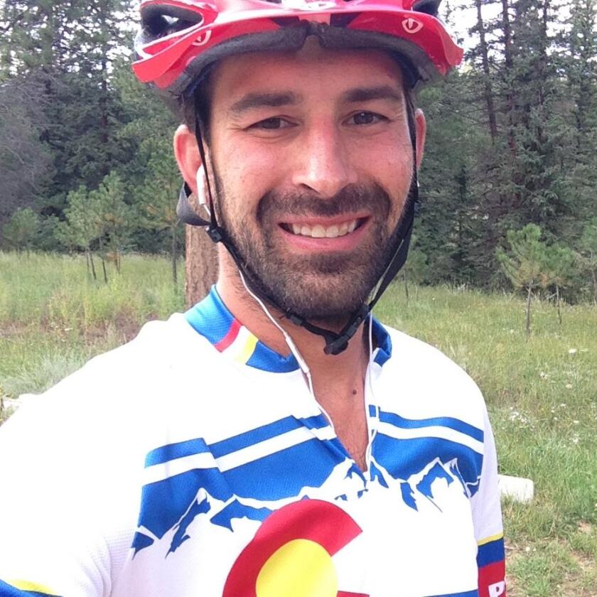 A man in a bike helmet and Colorado-themed bike shirt smiles in a selfie in front of a green tree line.