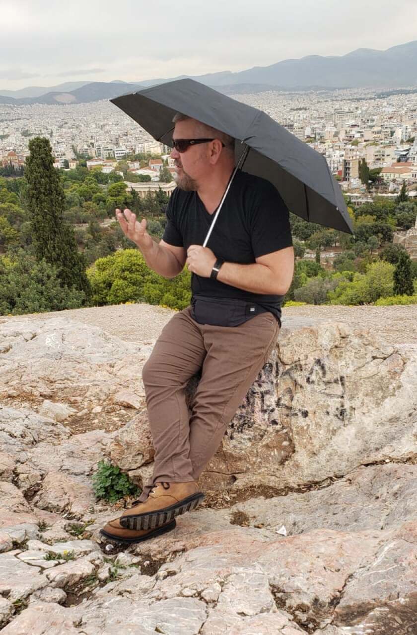 A man with a black umbrella reclines on a rock formation with Athens stretching away in the background.