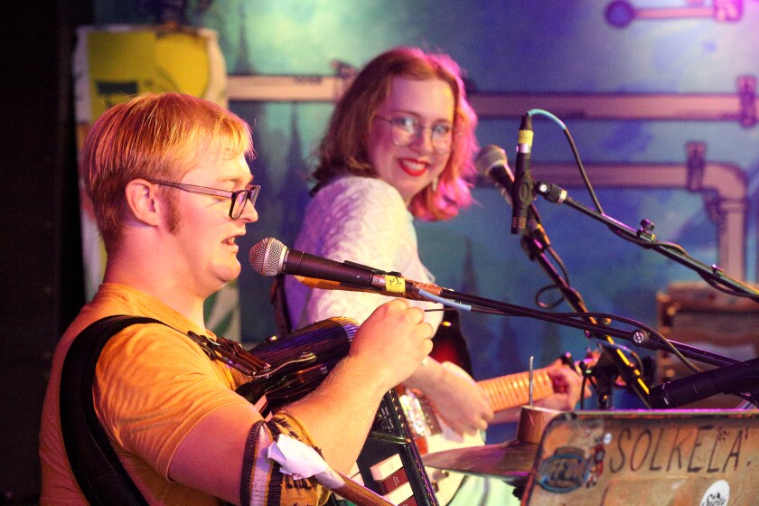 A man speaking into a microphone while performing on stage while a woman in the background looks on and smiles.
