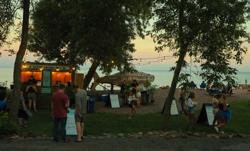 Exterior view of rocky lake beach with people gathered in small groups around a small kiosk selling drinks. Large wooden shark sits on shore in background.