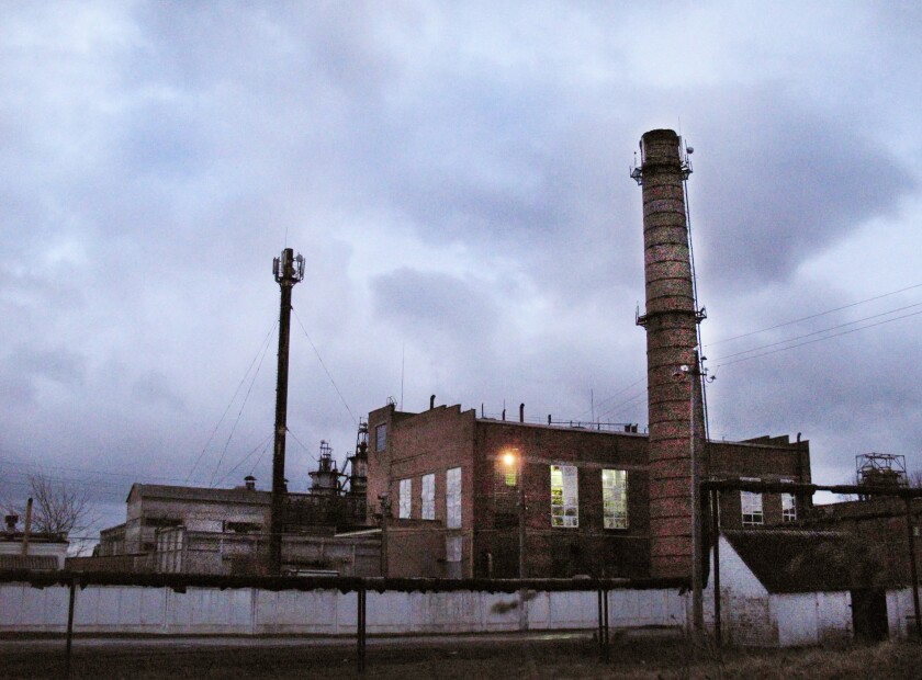 A relatively small sugar beet factory with a tall smoke stack stands against a dark, cloudy sky at dusk.
