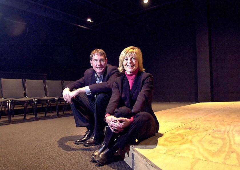 A light-skinned man and woman in business suits sit on a wooden stage in a dark room with theatrical seating visible in background.