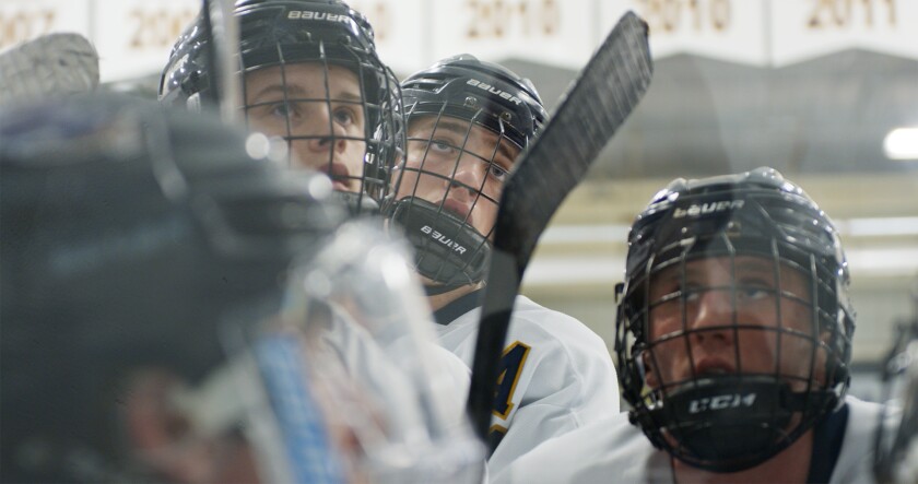 Teen hockey players, wearing white jerseys and black helmets, cluster together and wait expectantly.