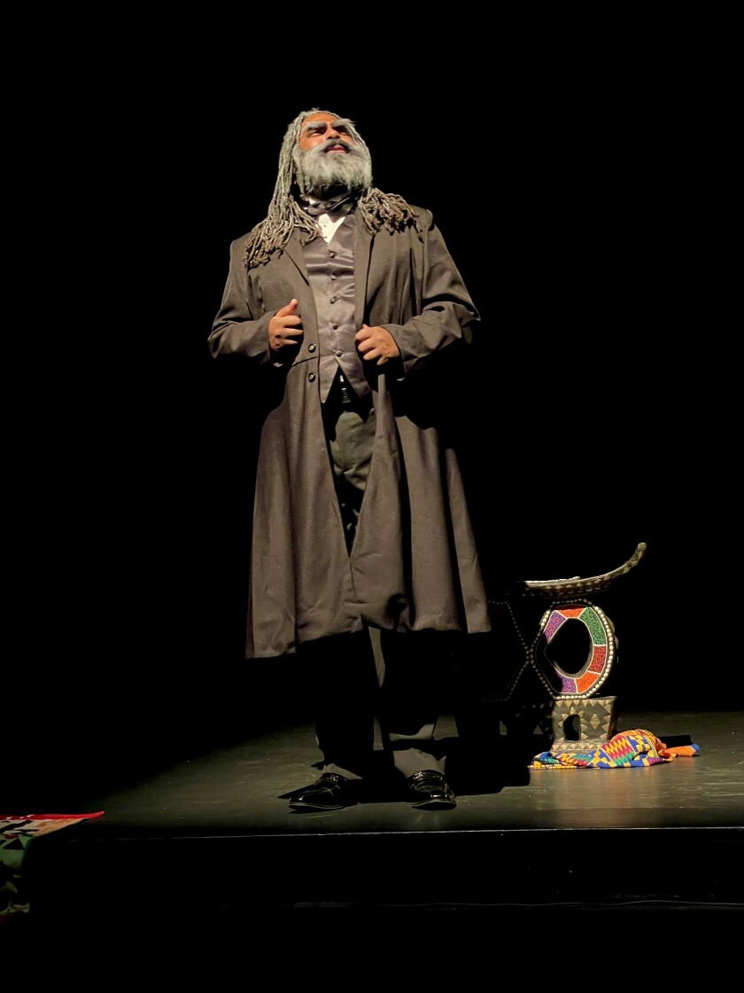 A Black man with long hair and a beard stands on a stage in the spotlight.