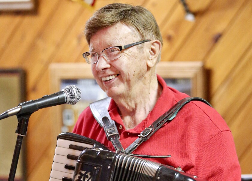 White man in his 80s smiles as he stands at a microphone wearing a red polo shirt and holding an accordion.