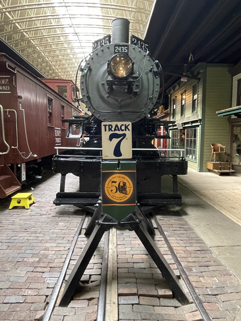 A black steam locomotive is seen behind a sign reading "TRACK 7" and a gold medallion banner with the Lake Superior Railroad Museum 50th anniversary logo.