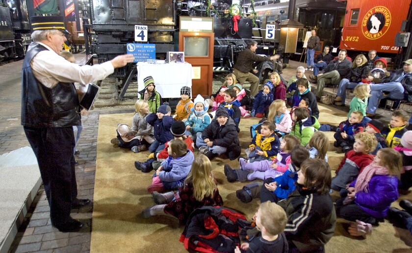 A large group of children in winter clothing gather on a carpet as a light-skinned man in a conductor's uniform talks to them.