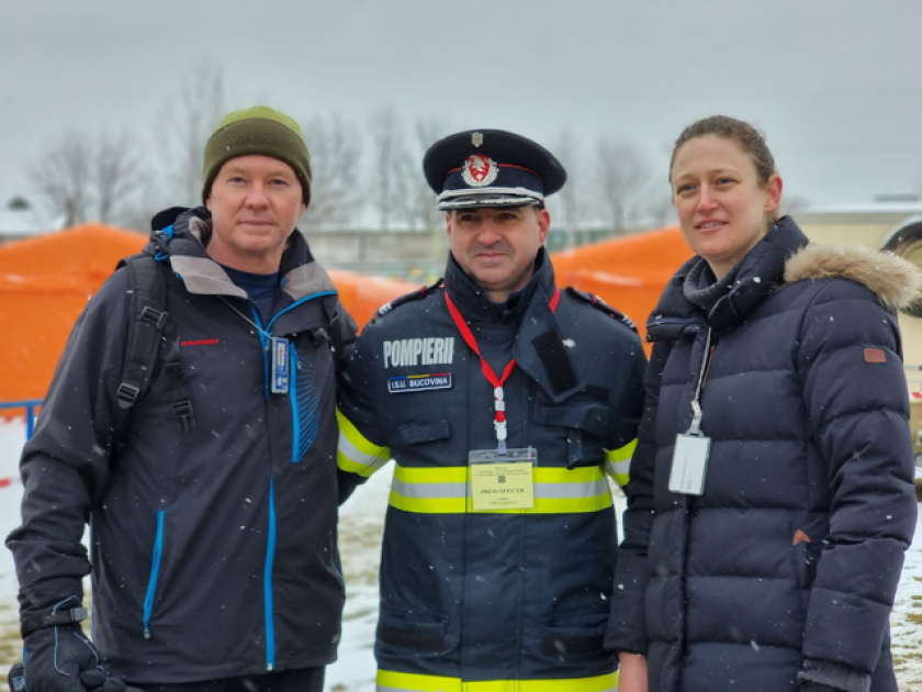 Standing outside in dark jackets while it snows, two men and a woman pose for a photo.