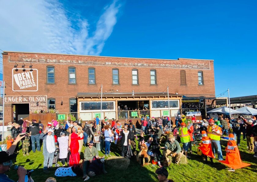 Large crowd of adults and children, many in costume, gather on a lawn outside a two-story brick building painted with the Bent Paddle Brewing Co. logo.