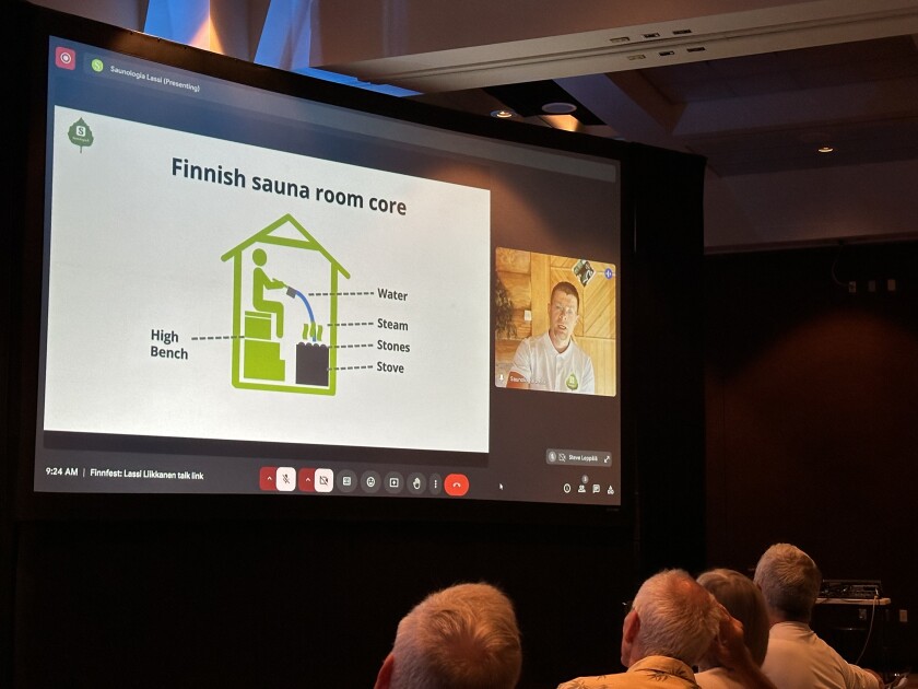 In darkened conference center, people sit watching a large screen. On the large screen, a small image of a white man's head is next to a large diagram labeled, "Finnish sauna room core."