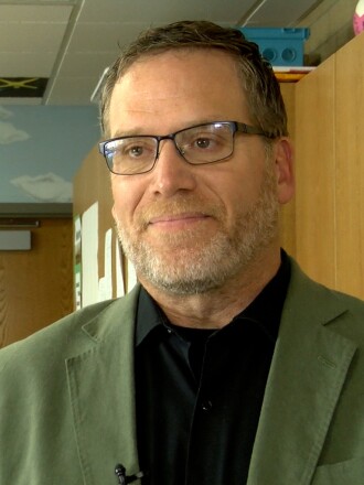 A man in an olive green sport coat, black shirt and glasses looks toward the camera.
