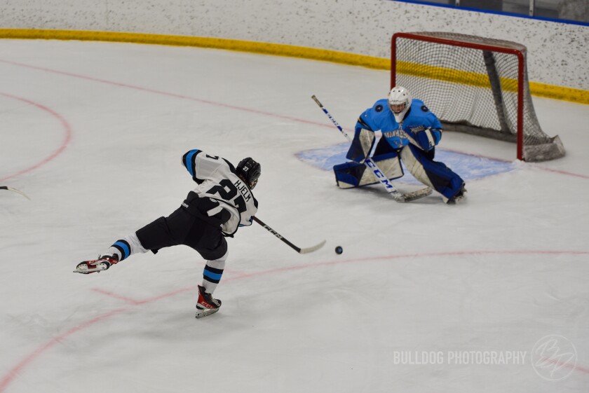 A player wearing a white jersey with blue and black trim shoots the puck at a goaltender wearing a light blue jersey with navy blue and black pads.