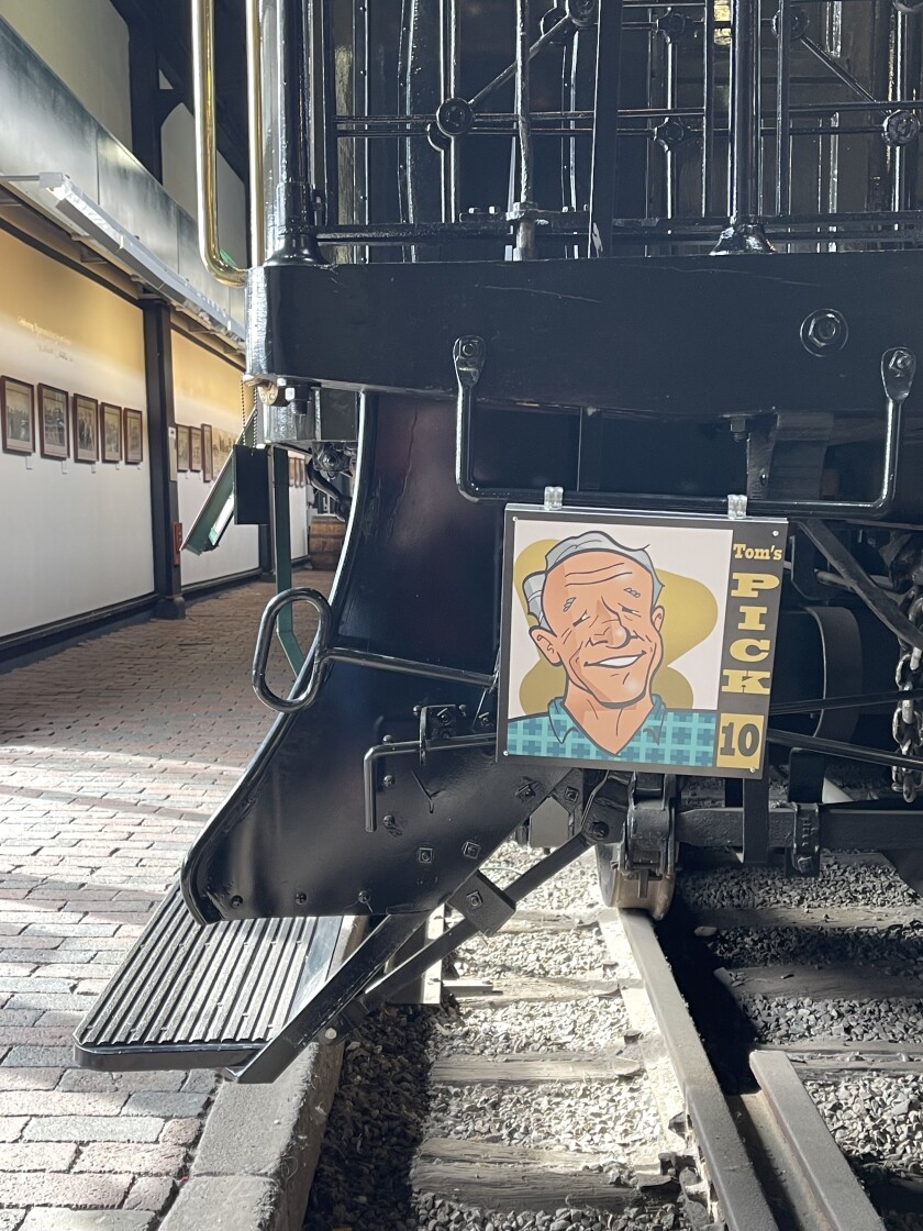 Close-up view of step on train car, with sign affixed reading "Tom's Pick 10." Sign features caricature of smiling grey-haired light-skinned man.
