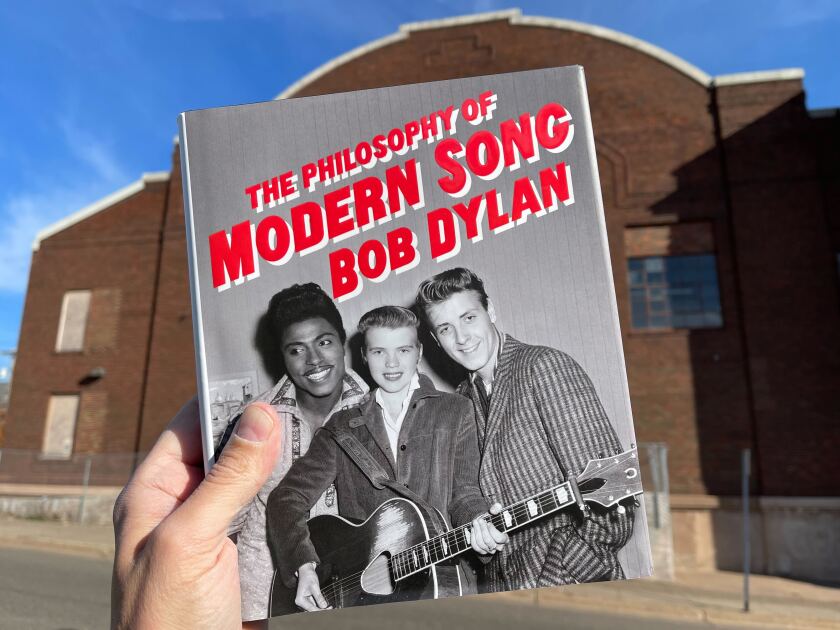 Hand holding book, Bob Dylan's "The Philosophy of Modern Song," in front of brick armory building. Book features cover photograph of three early rock and roll musicians posing together.