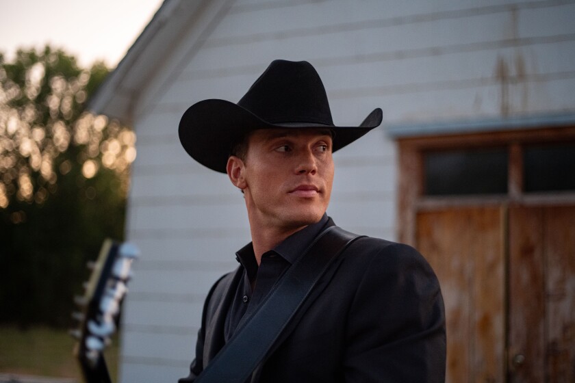 Light-skinned man wearing black suit and matching cowboy hat stands, looking reflective, in front of white-painted garage.