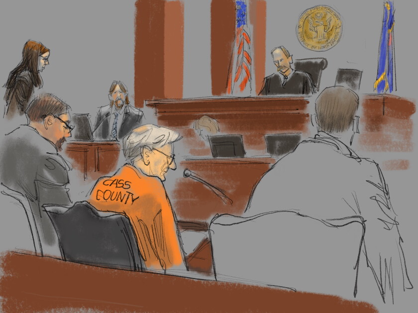 A scene inside a federal courtroom is seen in an illustration from the viewing area. At center is a white male judge presiding over the proceedings. Seated at a table surrounded by two suited men is an old white man in an orange prison jumpsuit which reads CASS COUNTY in black text. A white woman speaks a podium as well while another man is seen seated watching the proceedings.