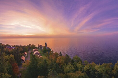 Sun rises over Lake Superior, with color fading from white to a purplish blue, in an aerial photo. Tree-lined shoreline with lighthouse seen in foreground.