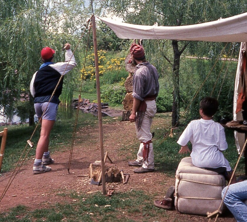 A teenage boy prepares to throw an ax at a wooden target in an outdoor area, as a costumed voyageur and a young boy look on.
