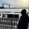 Man in black jacket and winter hat is seen from behind, looking over lake harbor with lift bridge in background. Railing stands behind him.