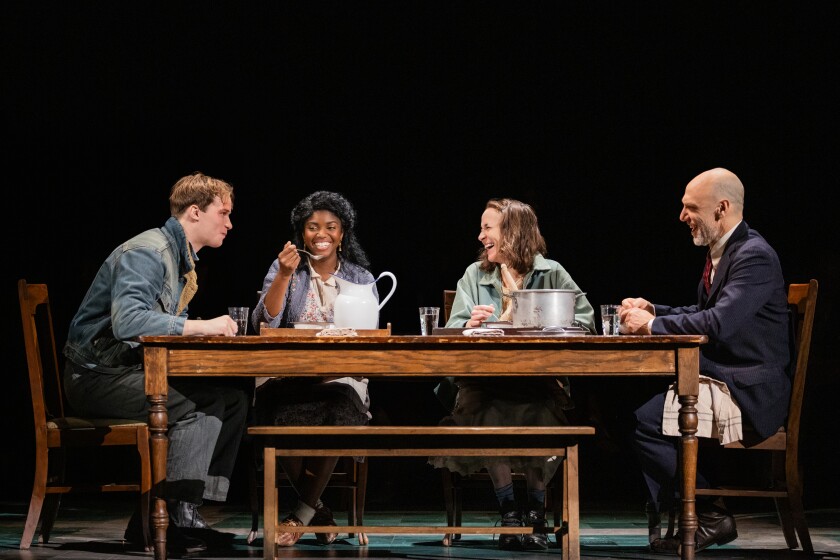 Four people dressed in 1930s garb sit around a simple wooden dinner table, sharing a meal. All are smiling and looking at one another.