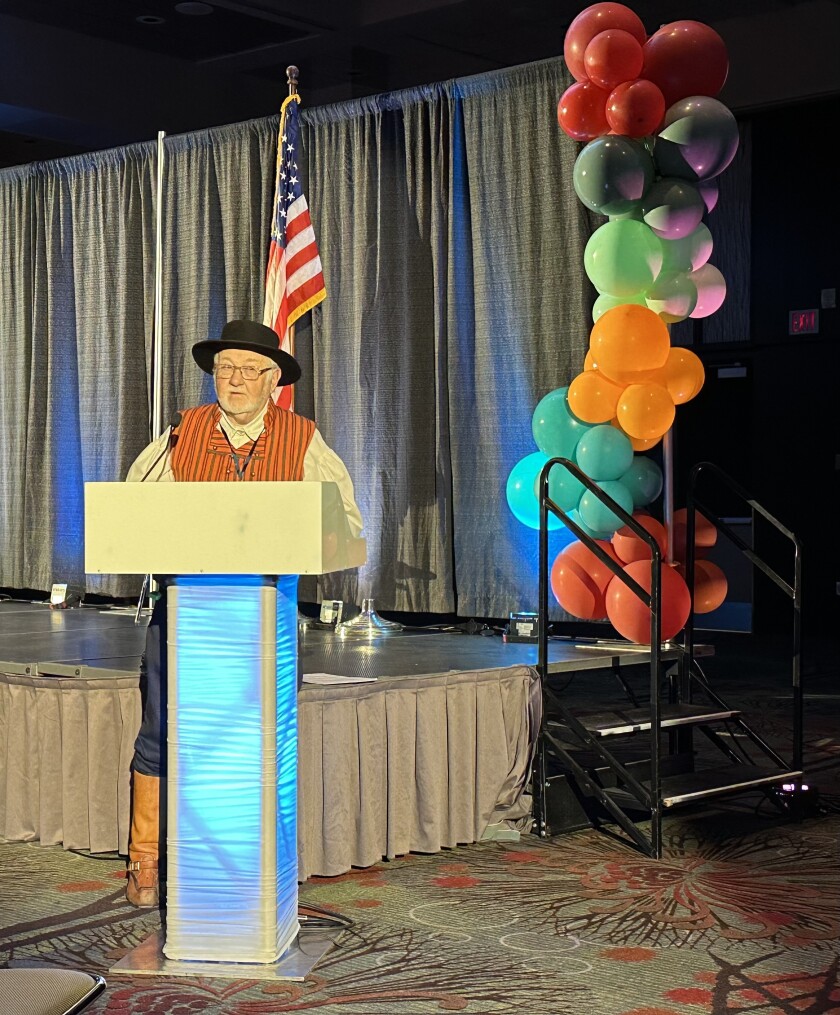 White man in his senior years stands at lectern in conference room. He wears knee-high tan boots, blue pants, white shirt with wide sleeves, striped red vest, and flat black hat with round brim.