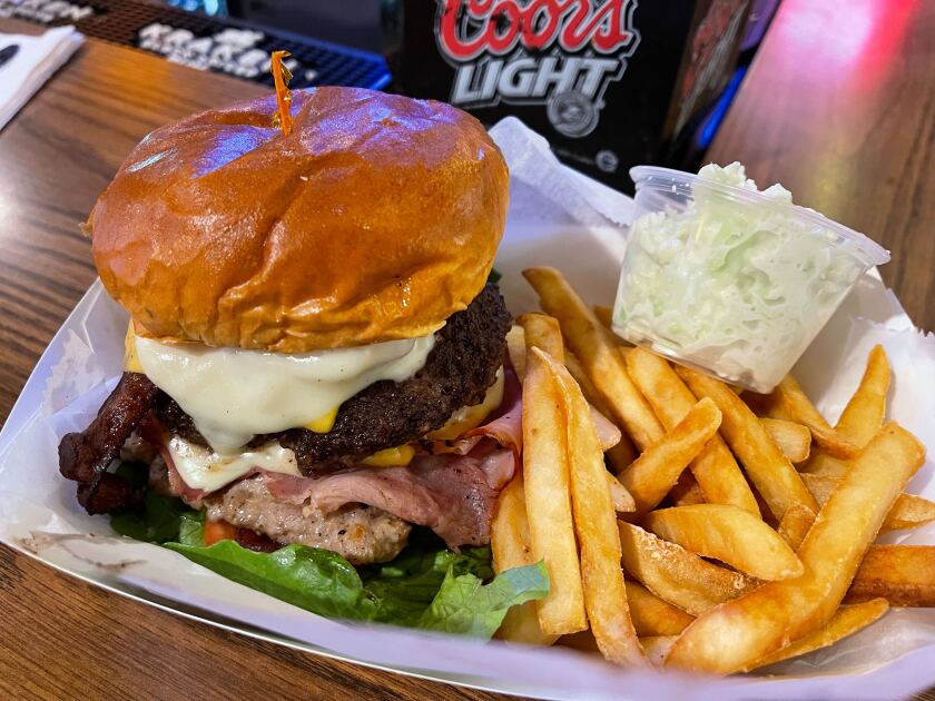 A very large hamburger stacked with other meats between the bun, in a basket with french fries and cole slaw.