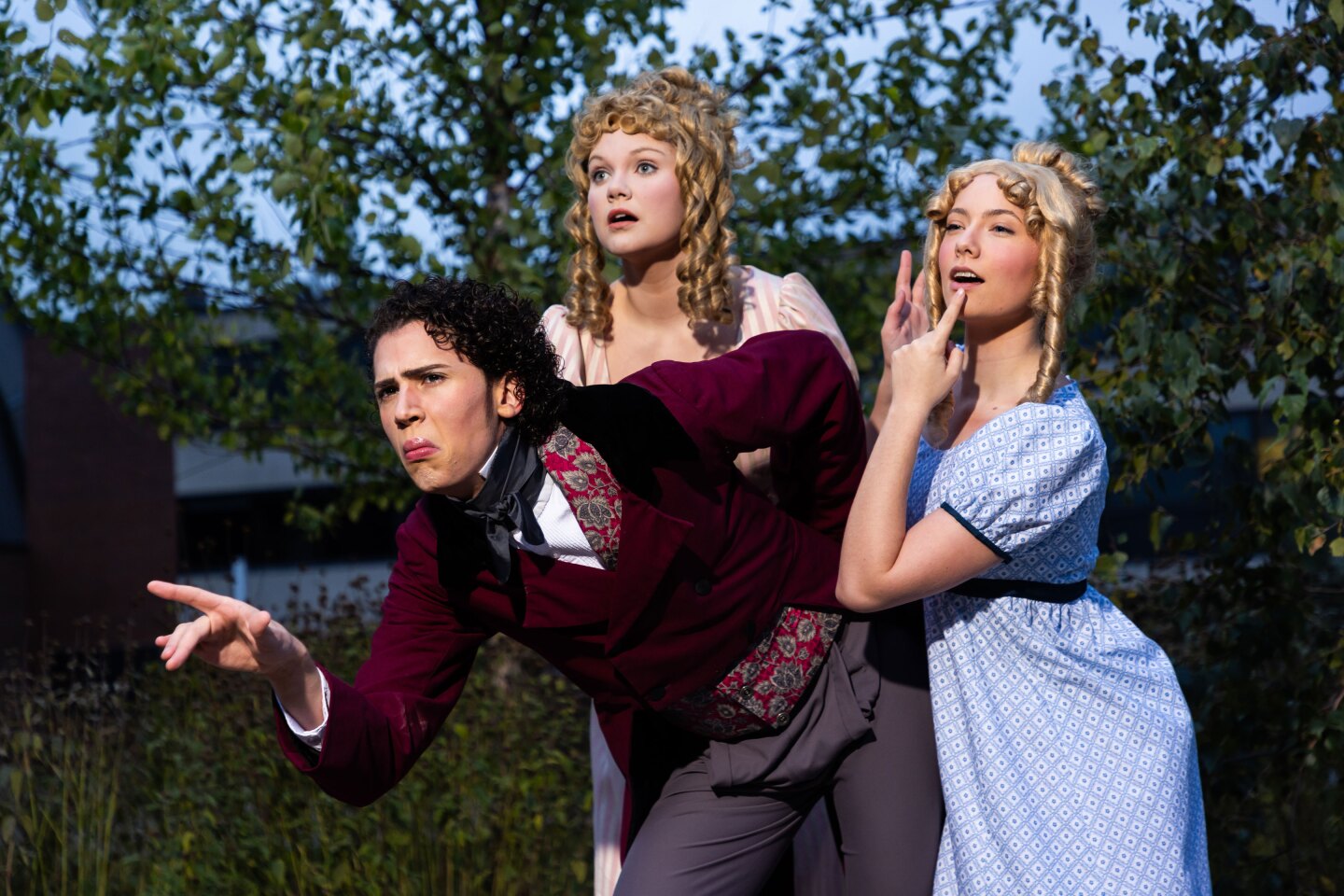Three light-skinned young adults in costumes evoking 19th century British gentry pose as if looking with great interest at something to the left.