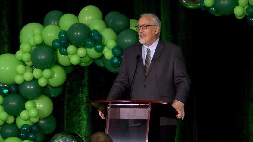 A man in a suit leans on a podium while speaking in front of a backdrop of green balloons.