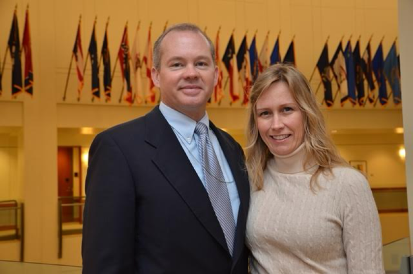 A man in a suit and a woman in a cream turtleneck smile together for a photo in a hall bedecked with many flags.