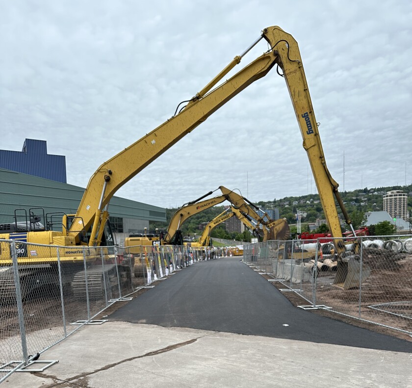 Three yellow excavator arms arc over paved pathway, with large blocky building at left and urban hillside in background.
