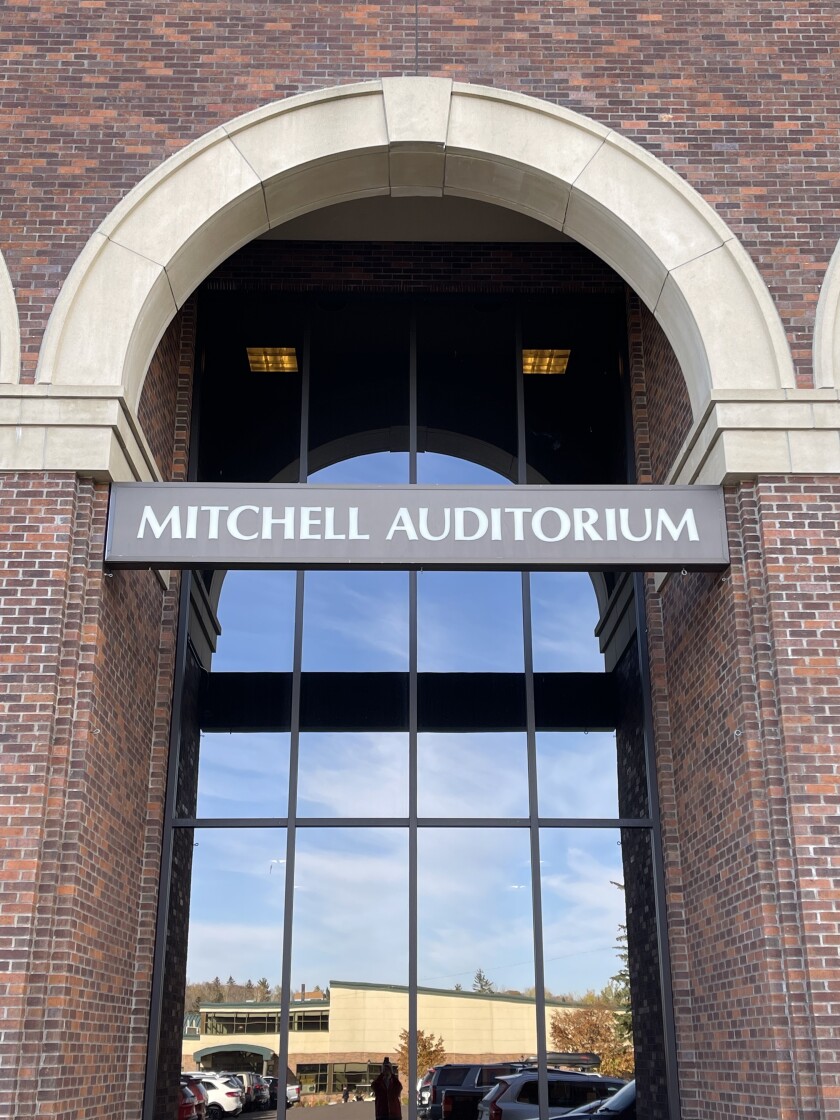 Exterior view of windows in a brick arch, with a sign reading MITCHELL AUDITORIUM spanning the arch. Reflection in windows shows blue sky over a low, large building.